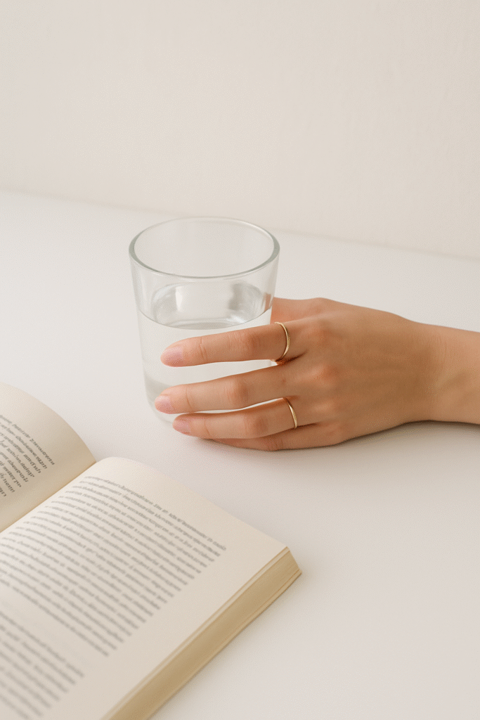 A minimal aesthetic scene with a slender hand wearing gold rings, gently holding a glass of water beside an open book on a white table, symbolizing clarity, focus, and mindfulness.